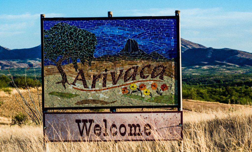 photo of mountain landscape with brown fields against green mountain with blue sky, centering a sign saying "Arivaca" and "Welcome" below it.